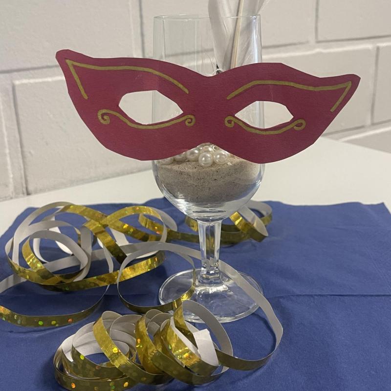 Glas mit Sand, weißer Feder und Perlen gefüllt mit Papiermaske als Tischdeko